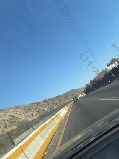 The image captures a roadside scene on a clear, sunny day, likely taken from a moving vehicle. The dominant features include a highway, a steep hillside, power lines, and a bright blue sky.

In the foreground, a multi-lane asphalt road stretches into the distance. It features distinct yellow lane markings. To the left of the driving lanes, a concrete barrier, painted with prominent yellow stripes, separates the road from a sloping, rocky hillside. Further down the road on the shoulder, a dark-colored vehicle, possibly a small truck or SUV, is visible. The reflection of power lines can be faintly seen on what appears to be the vehicle's side window from which the photo was taken, indicating the photo was shot through glass.

Adjacent to the road, on the left side, is a rugged and steep hillside. It appears arid, covered with dry, sparse vegetation, rocks, and some patches of reddish-brown earth.

Above the hillside and the road, a vast, clear blue sky dominates the upper portion of the image. Spanning across this blue sky are numerous sets of overhead power lines. These lines are supported by several tall, metal lattice power line towers (electricity pylons) that are visible on the hillside and further in the distance.

A white road sign with black text is visible in the upper-left area, partially obscured by the hillside's vegetation. The text on the sign reads: "REDUZCA SU VELOCIDAD," which translates from Spanish to "REDUCE YOUR SPEED."

The scene suggests travel on a highway in a semi-mountainous or hilly region, under bright daylight conditions.