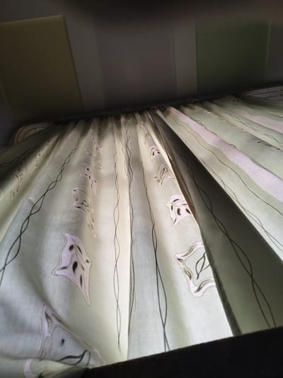 The image depicts a light green curtain with a repeating leaf pattern and thin black wavy lines. The curtain is illuminated from behind, creating a translucent effect. The top of the image shows a ceiling composed of panels, one of which is a lighter beige color. The bottom of the image shows a dark brown surface, possibly a shelf or windowsill. The scene appears to be indoors, with no visible people or explicit action. The image was taken in Melitopol, Ukraine.