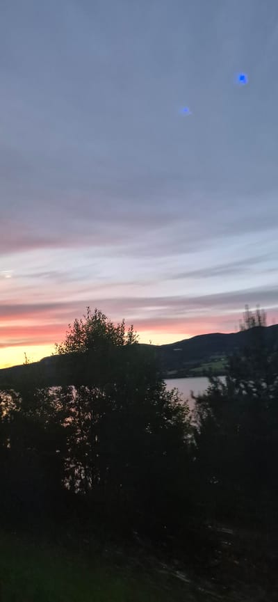 A wide-angle vertical shot captures a serene sunset over a body of water in Biri, Norway. The sky is a gradient of soft blues, purples, and pinks, with wispy clouds streaking across. Two small, bright blue rectangular lights are visible in the upper portion of the sky, their origin unclear. In the foreground, silhouetted trees obscure a view of a dark, calm lake and rolling hills in the distance. The overall atmosphere is peaceful and tranquil, illuminated by the fading light of day.