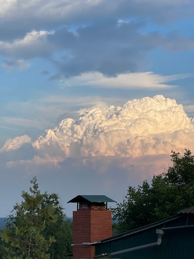 This image depicts a dramatic sky over a residential area. In the foreground, a brick chimney with a green metal cap stands prominently. To the right, a portion of a dark green roof with a gutter system is visible. Lush green trees, including a tall conifer on the left and denser foliage to the right, frame the scene. The background is dominated by a vast sky filled with large, textured cumulus clouds, illuminated by warm, golden light suggesting either sunrise or sunset. The lower portion of the sky features lighter blue hues interspersed with darker, wispy clouds. The overall impression is one of a tranquil, rural or suburban setting experiencing a moment of atmospheric beauty. The location is identified as Krasnokutsk, Ukraine. There are no people or animals present in the image.