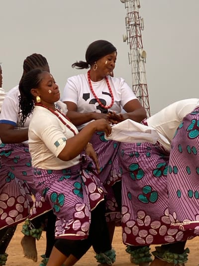 This vibrant photograph captures several women engaged in what appears to be a traditional cultural performance or ritual in Agunu, Nigeria. The women, with dark skin tones, are dressed in white short-sleeved tops and colorful, patterned ankle-length skirts or wraps. The skirts feature a dominant purple and maroon base with distinctive teal/green and light purple circular or shell-like motifs.
Each woman wears a long necklace made of bright red beads and prominent earrings, some hoop-shaped, others gold-toned drops. Their dark hair is styled in braids or ponytails. Notably, their faces are adorned with white markings, possibly traditional paint or chalk, on their cheeks and foreheads, lending a ceremonial aspect to their appearance. Their expressions are serene and focused, suggesting deep engagement in the activity. Green, leafy or fabric adornments are visible around the ankles of some participants, commonly used in traditional dances.
The setting is outdoors, under an overcast sky which provides soft, diffused lighting, indicating daytime. In the background, a large metal lattice structure, resembling a telecommunication mast or transmission tower, stands against the light sky. A partially visible black design, possibly a logo or symbol, is seen on the white t-shirt of one of the women. The overall scene conveys a moment of cultural celebration or ritual performance.