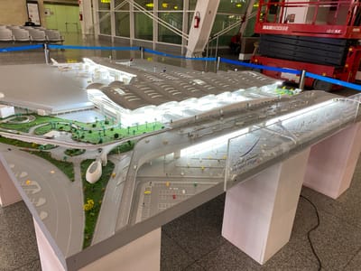 A detailed architectural model of Algiers Houari Boumediene Airport, located in Dar El Beïda, Algeria, is displayed. The model accurately replicates the airport's terminal buildings, runways, taxiways, and surrounding landscape, complete with miniature aircraft, vehicles, and trees. The model is illuminated, highlighting the intricate design of the terminal's roof structure and the interior spaces. In the background, a red scissor lift is visible, suggesting ongoing construction or maintenance work at the actual airport. Several individuals are present in the background, some observing the model and others engaged in work-related activities. The overall environment appears to be an indoor exhibition or display area, with flooring and lighting consistent with such a venue.