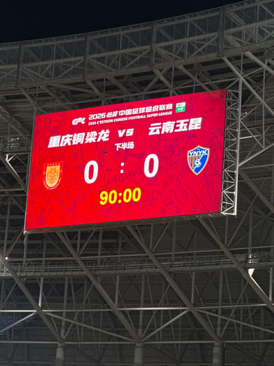 The image shows a brightly lit scoreboard hanging from the ceiling structure of a stadium. The scoreboard is red and displays the score of a football (soccer) match between Chongqing Tongliang Long and Yunnan Yukun, both teams with a score of 0. The clock shows 90:00 minutes, likely indicating the end of the match. The scoreboard also displays the logos of the two teams and information about the league. The stadium’s ceiling structure is visible, consisting of a complex network of metal beams. The location is identified by the team names as Longxing, China.