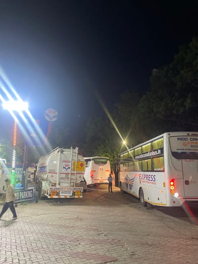 At night in Kajheri, India, a refueling station is depicted with multiple vehicles. An HP petrol pump sign is visible, illuminated by bright lights. A tanker truck with "CAPACITY 29000-LTRS" and "HP" branding is parked, presumably for refueling. To the right, two white passenger buses are present. The first bus has "FLIXBUS" branding, and the second bus features "INDO CANADIAN EXPRESS LUXURY INTERCITY SERVICE" and "www.indocanadianbus.in" on its side. A person in dark pants and a light-colored shirt is walking across the paved ground. Another person in a dark uniform is standing near the second bus. The environment is characterized by paved ground, trees in the background, and artificial lighting from the station and vehicle lights. A "NO PARKING" sign is also visible.