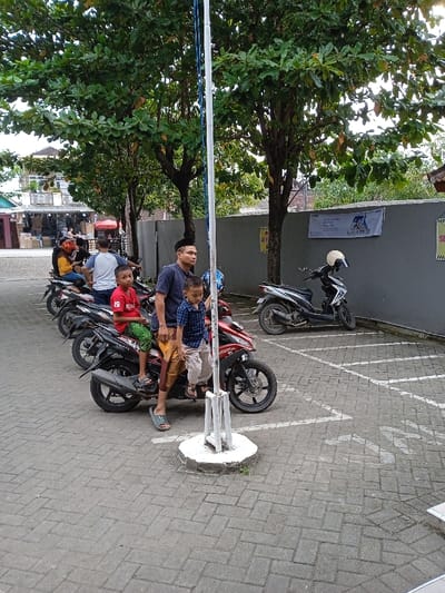 An outdoor scene in Sarimulyo, Indonesia, depicts a paved area, likely a parking lot, featuring three individuals and multiple motorcycles. In the immediate foreground, a white pole on a circular concrete base stands vertically. To the left of this pole, an adult male and two young boys are positioned around a red and black scooter-style motorcycle.
The adult male, wearing a dark blue t-shirt, light brown sarong, light blue slide sandals, and a dark peci cap, stands on the left side of the motorcycle, his left hand on the handlebar. He looks towards the right. An older boy, wearing a red t-shirt with "19" and green patterned shorts, sits on the passenger seat of the same motorcycle, facing forward. A younger boy, clad in a blue and white checkered shirt and light shorts, sits on the motorcycle seat in front of the adult male; his right hand is gripping the central white pole.
The ground is covered with grey interlocking bricks arranged in a herringbone pattern, with white parking space lines visible on the right side. Several trees with dense green foliage are distributed across the background, providing shade. To the left, additional motorcycles are parked, with individuals visible on some. In the distant left background, a multi-story building with balconies and a commercial area at its base can be seen. To the right, a plain grey wall serves as a backdrop, against which two scooters are parked, one silver and one black, the latter with a white helmet resting on its handlebar. A white banner with blue text and a graphic is affixed to this wall, above a partially visible yellow sign. The scene is illuminated by natural daylight.