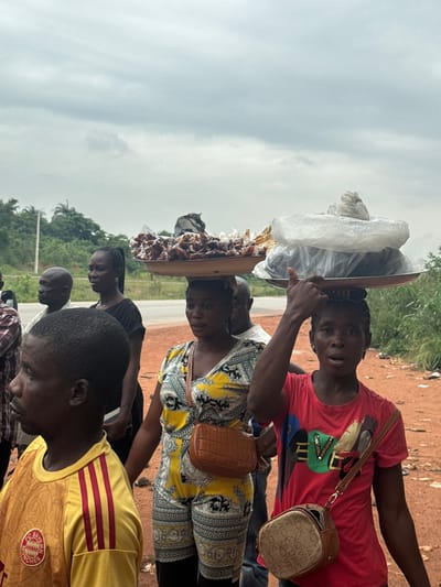 This media file depicts a street scene in Umuoye, Nigeria. The foreground features several people, including a young man wearing a yellow shirt with red stripes and a logo that appears to read "FC BAYERN MUNCHEN." To his left, another person is partially visible. In the middle ground, a woman with braided hair is seen from the shoulders up, looking down. Behind her, another woman wearing a bright yellow and black patterned outfit is shown from the chest up, with a small brown purse hanging from her shoulder. She is looking towards the camera. Dominating the upper portion of the image is a woman balancing a large tray of goods on her head. The tray contains items wrapped in white plastic and what appears to be food items, possibly dried fruits or snacks. She is looking directly at the camera with an open mouth, suggesting she might be speaking or reacting to the photographer. The background shows a reddish-brown dirt path and a lush green area with trees, indicative of a rural or semi-urban setting. The sky is overcast, suggesting a cloudy day. The overall impression is a candid snapshot of daily life and commerce on a Nigerian street. There is no clear indication of the time of day, but the lighting suggests it is daytime. The emotions displayed by the subjects appear natural and unposed, with some individuals looking directly at the camera.