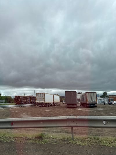 This is a ground-level, eye-level, vertical, wide shot of a logging area and truck yard under a cloudy sky. In the foreground, a metal guardrail runs horizontally across the bottom of the frame, separating the viewer from a muddy, dirt area with patches of grass. Behind the guardrail, several large trucks are parked. The leftmost truck is a logging truck, carrying a large load of cut logs secured with blue straps. Next to it is a white semi-trailer truck. Further to the right are two more semi-trailer trucks, one a dark color and the other white. In the background, there is a small building with "ПРОДУКТЫ" (Products) signage, a few parked cars, and trees. The ground is wet and appears to be a mix of mud and dirt, with tire tracks visible. The sky is overcast with heavy, dark clouds. The scene suggests a commercial or industrial area, likely a log processing or distribution center, in Alnashi, Russia. There are no people actively engaged in actions.
