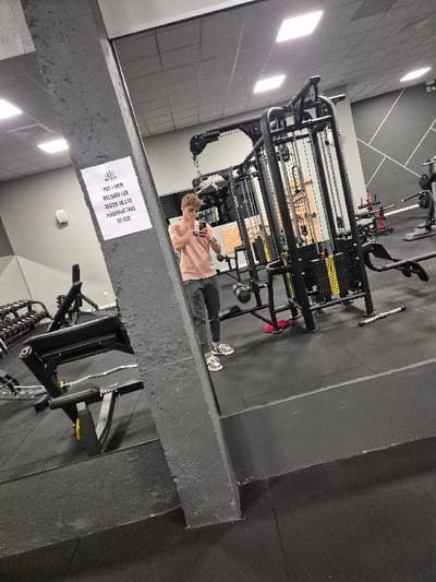 The image depicts the interior of a modern gym located in San Lorenzo de El Escorial, Spain. A young male, reflected in a large mirror, is captured in a selfie. He wears a light peach-colored short-sleeved t-shirt, dark trousers, and white athletic shoes with black accents. His right hand holds a smartphone positioned towards his face, while his left hand rests near his mouth/nose. He is looking directly at his reflection/the camera.

The gym features dark grey rubber flooring and a grey suspended ceiling with multiple square LED light fixtures. The walls in the background have a geometric pattern of dark and light grey sections. Prominently visible in the mirror's reflection are various pieces of gym equipment, including a black multi-station cable machine with yellow weight stacks in the mid-ground and a black weight bench with a barbell in the left mid-ground. Other gym apparatus, some with red components, are partially visible in the background.

A textured, dark grey concrete pillar extends vertically through the left foreground, partially obscuring the reflection. Affixed to this pillar is a white rectangular sign displaying text in Spanish: "PROHIBIDO DEJAR LOS BARRAS DE LAS MÁQUINAS TRAS SU USO" (Forbidden to leave the bars from the machines after their use).