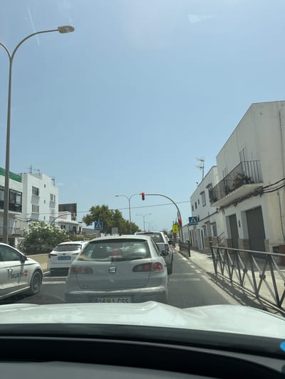 The image depicts a street scene in Can Negre, Spain, on a bright, sunny day. The perspective is from inside a vehicle, looking forward through its windshield, which shows reflections. The street is lined with white buildings, some with balconies and potted plants. There is traffic congestion, with several cars stopped in a lane. A white car with the "WURTH" logo is visible on the left. A light gray hatchback with the license plate "0691 CSS" is directly in front, and a white car with license plate "2334 MMX" is ahead of that. Traffic signals, including a red light, are visible. Pedestrian crossing signs are also present. Trees and vegetation are present along the street.
