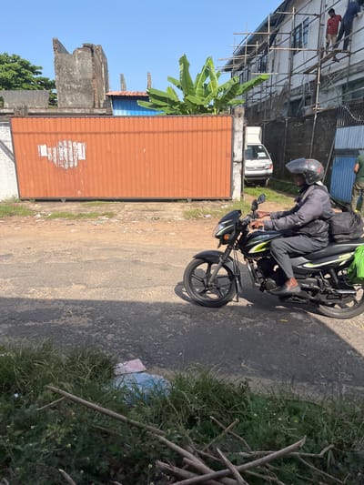 The image depicts a street scene in Moratuwa, Sri Lanka. A man wearing a helmet and a gray jacket is riding a black motorcycle. The motorcycle has green and yellow accents. He is carrying a black backpack on his back and a green bag is attached to the rear of the motorcycle. The man is looking to his right. In the background, there is an orange corrugated metal gate. To the left of the gate, a damaged concrete structure is visible. Further in the background, a white van is parked. A building with scaffolding is under construction on the right side of the image, and two men are working on it. The ground is a mixture of asphalt and dirt, with some patches of grass and debris. The sky is clear and blue.