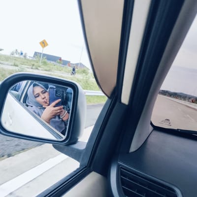 The image is a unique selfie taken from inside a car, focusing on a woman reflected in the vehicle's side mirror. The woman, wearing a light grey hijab, is holding a purple smartphone and appears to be taking her own picture, posing with slightly puckered lips. She wears a gold ring on her right ring finger and multiple bracelets, including pearl-like ones, on her right wrist.
The setting is an outdoor roadside environment during what appears to be an overcast day. Through the side mirror and the car's windows, a paved road with white lane markings and a silver guardrail is visible. In the background to the left, a yellow diamond-shaped road sign with a black curved arrow points left, indicating a bend in the road. Beyond the sign, green foliage, possibly a hillside, and several buildings with dark roofs are present. A person on a motorcycle can also be seen further down the road. To the right, through the front passenger window, an expansive road stretches into the distance with orange and black chevron signs marking a curve, and faint outlines of mountains or hills are visible against the sky.
The car's interior, including parts of the door panel, window frame, and dashboard with an air vent, frames the scene. On the bottom right of the car's passenger side window, text such as "DA", "S02000 - B", and "E1 43R - 000205" is visible, likely product or safety information. The image captures a personal moment of photography while traveling in Cotkareung, Indonesia.