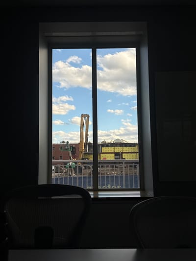 The image captures a construction and demolition site in Waltham, United States, viewed through a rectangular, vertically divided window from a dimly lit interior. In the immediate foreground, two dark-silhouetted Herman Miller Aeron office chairs and a table surface are visible, set against dark grey walls with a partially visible white board on the right.
The exterior scene, illuminated by a bright blue sky with scattered white cumulus clouds, features a large yellow high-reach demolition excavator with an extended boom prominently centered. To the left, a green aerial work platform is positioned near a multi-story red brick building. To the right, a structure partially covered in yellow insulation panels is undergoing construction or demolition. The ground area of the site exhibits disturbed earth and debris. A white metal railing separates the immediate foreground from the main construction zone, beneath which a blue banner with partially visible white text ("ctor") is present.