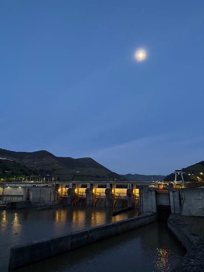 The image captures the Bagaúste Dam on the Douro River, located near Lamego, Portugal, during twilight or night conditions. The upper half of the frame displays a dark blue sky, featuring a distinct, luminous full moon with a visible halo in the upper right quadrant. The horizon is defined by dark, undulating hills or mountains, showing some terraced or cultivated sections. The central and lower portions of the scene are dominated by the concrete dam structure. Multiple large sluice gates are visible, fronted by dark grey concrete pillars. The water flow areas immediately behind these gates are strongly illuminated by warm, yellow artificial lights, casting pronounced reflections on the dark, placid river water. A thick, dark grey concrete barrier or wall extends horizontally across the mid-ground water surface. To the far right, a tall, white gantry-like structure with bright white lights is present, positioned adjacent to a further section of the dam. No individuals are depicted, and the scene is static.