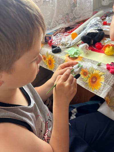 The image shows a close-up of a child's hands as they sculpt with light green clay. The child is wearing a grey tank top with black trim. The setting appears to be a home, with a tablecloth featuring a sunflower pattern visible. Various crafting supplies, including colorful clay and packaging, are scattered on the table. The lighting suggests it is daytime, with natural light illuminating the scene. The child's focused expression, visible in the cropped images, indicates engagement in the activity. The location context provided is Маркидоновка, Russia. No specific text is clearly legible in the image.
