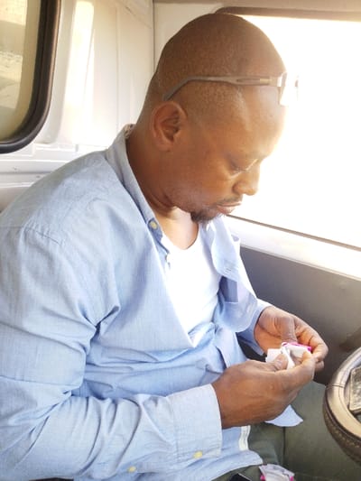 A middle-aged Black man sits indoors, possibly in a vehicle, wearing a blue button-down shirt over a white undershirt. He's holding a small, possibly medical, item wrapped in tissue. Sunglasses are perched on his head. He's looking down at the item in his hands with a focused expression. The background is blurred by strong sunlight, and only the vehicle interior is visible.