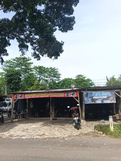 The image depicts a roadside motorcycle repair shop in Indonesia. The shop has a simple structure with a corrugated metal roof. Banners advertise services, including "Amrizal Service Jok Motor" for selling used spare parts and "Bengkel Motor" for tune-ups, service, and spare parts. Several motorcycles are parked around the shop, indicating active business. The surrounding environment includes trees and vegetation, suggesting a rural or suburban setting.
