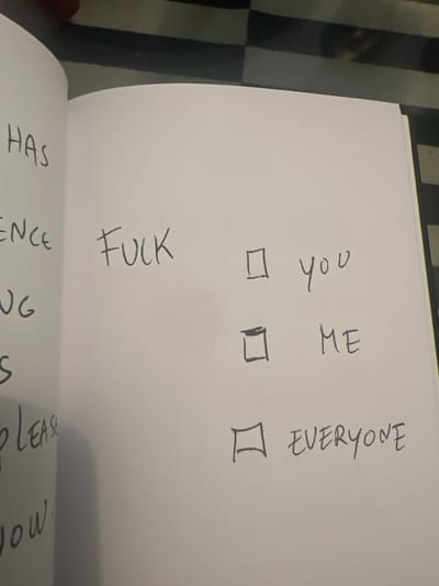 An open book displays handwritten text. The word "FUCK" is written vertically beside three checkboxes. Next to the checkboxes, also written vertically, are "You," "Me," and "Everyone." The lower edge of the image shows portions of other handwritten words, including "HAS," "ENCE," "ING," "S," "PLEASE," and "NOW." The background is a checkered surface.