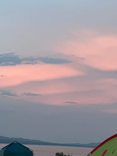 The image depicts a serene natural landscape with a touch of human presence. The sky is filled with soft, wispy clouds, tinged with hues of pink and blue, suggesting either sunrise or sunset. In the foreground, partially visible, are two camping tents. One is a vibrant green with red trim, and the other is a deep blue, both appearing to be set up for outdoor recreation.
The background features a body of water, likely a lake or river, with a distant shoreline characterized by rolling hills or mountains. The overall atmosphere is peaceful and tranquil, indicative of a quiet moment in nature. The time of day is either early morning or late evening, given the soft lighting and the colors in the sky. The weather appears calm. The location is identified as Kolodezny, Russia. There is no visible text in the image.
