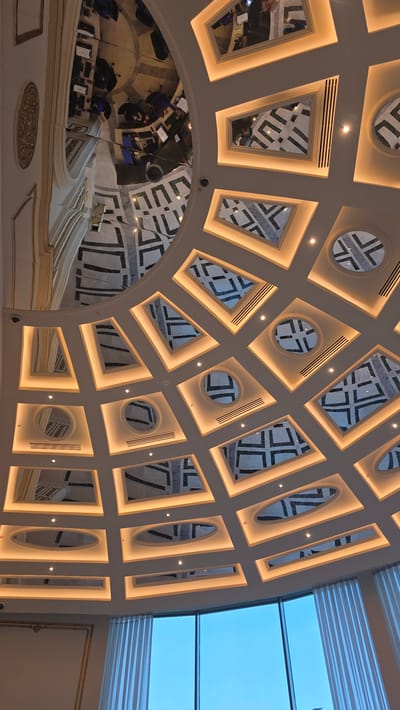 This is a low-angle, interior shot of a domed ceiling with recessed lighting and mirrored panels. The ceiling has a complex geometric pattern, with square and rectangular panels containing black and white designs, possibly marble. Some circular panels are also present. The lighting creates a warm, ambient glow around the edges of the panels.

In the mirrored panels, reflections show a modern interior with several people seated at desks with laptops. The reflections suggest a professional or office environment. The ceiling is part of a larger architectural structure, with ornate molding visible in some areas. Below the ceiling, large windows offer a view of a bright blue sky, indicating daytime. The overall impression is one of modern, sophisticated interior design. There is no information that places this scene specifically in Agios Epiktitos, Northern Cyprus / Cyprus.