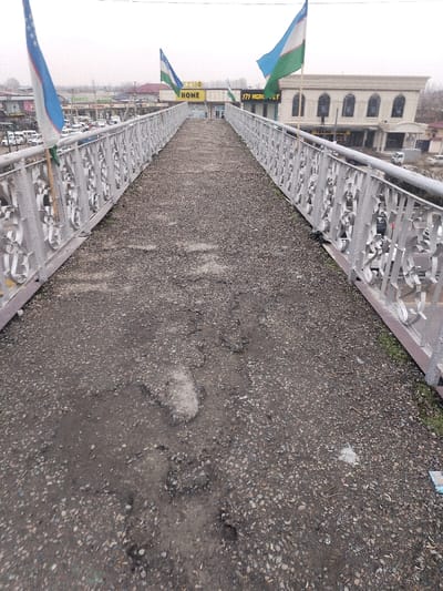 An elevated pedestrian overpass, situated in the city of Tashlak, Uzbekistan, is depicted from a low-angle perspective, looking northeast down its length. The overpass pavement is constructed of a dark, worn aggregate material, exhibiting significant degradation with numerous potholes, cracks, and uneven surfaces, particularly prominent in the foreground. Silver-colored metal railings with ornate, cutout scrollwork patterns line both sides of the walkway. Several flags of Uzbekistan, characterized by blue, white, and green horizontal stripes with a red border and a white crescent moon and stars, are mounted along the railings and in the background.
To the left, below the overpass, a street or parking area contains multiple vehicles, primarily white vans and cars, indicative of a commercial zone. Buildings with various signage, including one partially visible as "MARKET," are present beyond the vehicles. To the right, below the overpass, a light-colored, multi-story building with arched windows and decorative facade elements stands adjacent to a street. Further in the background, large signs identifying "TEHNO HOME" and "777 AGRO" are visible on commercial structures. The sky is overcast, and sparse green vegetation is present at the base of the railings where they meet the overpass surface. No individuals are visible on the overpass.