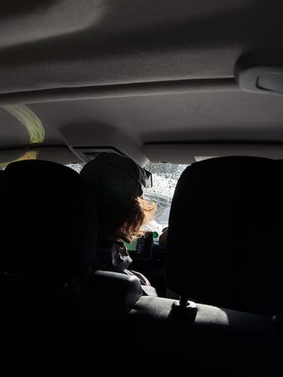 A person with reddish-brown hair and a gray hat appears to be sleeping in the back of a vehicle, with their head resting against the window. The window has a speckled appearance, possibly due to water droplets. The vehicle interior consists of a light gray wall and dark seats. The lighting is uneven, with bright sunlight filtering through the window and casting shadows. No specific details indicate the location is Saint-André, France.