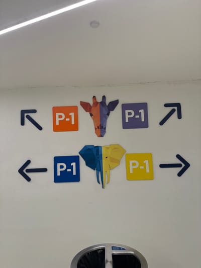The image shows a section of a white wall with various directional signs and colorful geometric animal depictions. Two dark blue arrows point upwards, one diagonally left and the other straight up, indicating directions. Below these, there are four square signs, each displaying "P-1" in white text. Two signs are purple and yellow, positioned next to each other, with the purple sign on the left and the yellow sign on the right. Below these, there are two more "P-1" signs, one orange and one blue, also side-by-side, with the orange on the left and the blue on the right.
Between the upper and lower sets of "P-1" signs are two stylized, polygonal animal figures. The left figure is predominantly purple and orange, resembling a bird or dragon in flight. The right figure is yellow and blue, appearing to be the head of a similar mythical creature or perhaps a stylized dinosaur.
Below the lower "P-1" signs, two more dark blue arrows point downwards, one diagonally left and the other straight down.
To the far right of the image, a silver and grey trash can with a black liner is partially visible. The wall itself appears to be clean and well-lit. The overall impression is of a well-marked hallway or public space, possibly in a modern building or exhibition. The geometric animal art adds a whimsical or educational element to the directional signage. The lighting suggests an indoor environment, likely during the day. There are no people visible in the image.