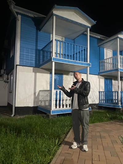A man stands on a wooden walkway next to two blue and white two-story buildings. The buildings are similar in design, with railings and paneled walls. The man is bald, wearing a black leather jacket, a white shirt, and gray pants. He is gesturing with his hands. The walkway is made of rectangular wooden blocks. There is grass growing next to the walkway. The background is dark, possibly night time. The location is inferred to be in Samarskoye, Russia.