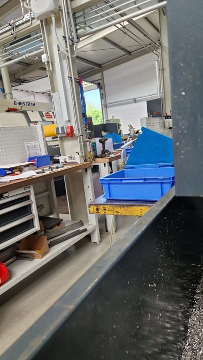This media file depicts an indoor industrial or workshop setting, likely in Ichenhausen, Germany, given the location context. The image is oriented vertically, with the scene appearing upside down from a standard perspective.

The primary subjects are various pieces of machinery and industrial equipment. Prominently featured is a large blue plastic bin situated on a metal frame. To the left and slightly below the blue bin, there's a piece of machinery with green and black components, possibly a manufacturing or processing machine. Further to the left, a complex network of pipes and conduits runs along the ceiling and walls, suggesting an active operational environment. There are also some workbenches with tools and supplies visible.

In the foreground, a dark, textured surface, possibly a table or machine part, occupies the bottom portion of the image, with what appears to be metal shavings or debris scattered across it. The background shows a bright, open area with natural light, hinting at a window or open doorway revealing trees and a blue sky, indicating it is daytime with fair weather.

Several labels and markings are visible. On a light-colored vertical support structure, the text "B 465 T2 Y2" is clearly legible. There are also smaller labels and signs on various pieces of equipment, though they are not clearly decipherable.

The scene appears to be a functional workspace, possibly a factory floor or a well-equipped workshop. There are no people visible in the image, suggesting the activity, if any, is either automated or occurring off-camera. The overall impression is one of an organized industrial environment.