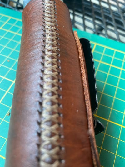 The image shows a close-up of a brown leather item, possibly a sheath or case, with decorative stitching. The stitching is a light tan color and forms a criss-cross pattern along the edge. The leather appears textured and slightly worn. Part of a black metal object is visible inserted into the leather item. The background consists of a green cutting mat with yellow grid lines, and a blurred black mesh structure. There are no people or animals visible in the image. The image does not provide any information that could be used to determine its location in the city of Lütjenwestedt, Germany.