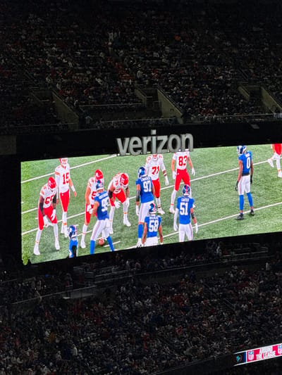 The image depicts a large scoreboard displaying an American football game at MetLife Stadium in East Rutherford, United States, likely during an evening event due to the dim stadium lighting. The scoreboard prominently features a "verizon" logo above the active game footage. On the screen, two teams are shown on a green artificial turf field: one in red and white uniforms (Kansas City Chiefs, with players such as #15, #57, #71, #83 visible) and the other in blue and white uniforms (New York Giants, with players #0, #93, #97 [Lawrence], #58 [Okereke], #51 [Carter] visible). Players are lined up on the field, with a football positioned on the ground near a crouched blue player, suggesting an active play. Below and to the sides of the scoreboard, large sections of stadium seating are visible, filled with numerous spectators. In the bottom right corner of the overall stadium view, a smaller graphic reads "AROUND THE LEAGUE" with NFL logos.