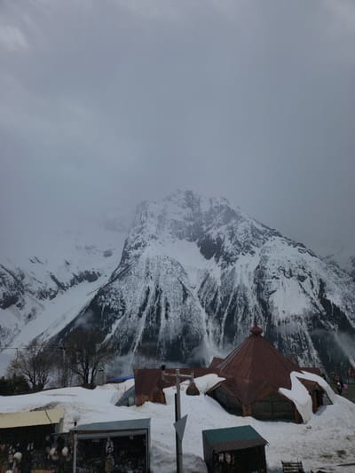 The image shows a snowy mountain landscape in Dombai, Russia. The left side of the frame is filled with a dense, grey fog obscuring the base of the mountain. Further up, the mountain's rocky face is visible, covered in snow, with dark streaks of exposed rock running through the snow-covered areas.  

To the right of the mountain, a cluster of buildings stands on the snow-covered ground. These buildings have reddish-brown roofs with snow piled on top. There appears to be a building with a grey corrugated metal roof. A few bare trees are visible between the mountain and the buildings. Faint, blurred figures of people can be seen at the bottom right of the frame.