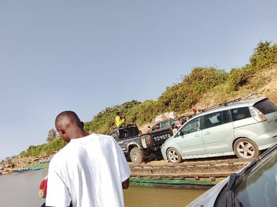 A daylight scene depicts a river crossing in Ibi, Nigeria. The foreground is dominated by the back of an adult male, bald or with very short hair, wearing a white short-sleeved t-shirt, looking forward towards the river and bank. Vehicles are positioned on a wooden ferry platform, which is floating on a calm, brown body of water.
From right to left on the ferry, a light green or silver minivan (possibly a Mazda) with roof racks is visible from its rear-right side. Behind it and slightly to the left, a black Toyota pickup truck with "TOYOTA" visible on its tailgate carries at least one individual in a yellow shirt in its truck bed, near the cab. Other less distinct vehicles are visible further back on the ferry. The front portion of a vehicle, likely the one from which the photograph was taken, is visible in the bottom right corner.
To the left of the ferry, a river bank features numerous small, brightly colored boats (blue, yellow, green) pulled ashore. To the right and extending into the background, a steep, vegetated incline is covered with dense green and brown scrub brush. The sky is clear and light blue, devoid of clouds.