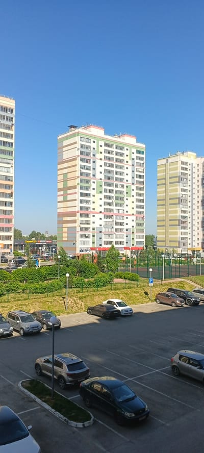 The image depicts a sunny day in Zonalnaya Stantsiya, Russia, showcasing a residential area with modern multi-story buildings and a parking lot filled with cars. In the foreground, a paved parking lot occupies the lower half of the frame, with several parked vehicles including a silver SUV, a dark sedan, a white sedan, and a silver hatchback. A street light stands in the center of a small, landscaped island within the parking lot.

Behind the parking lot, a grassy embankment slopes upwards, leading to a green recreational area. This area features a playground with a slide and climbing structures, as well as a sports court with a green surface and what appear to be basketball hoops. Trees and bushes are interspersed throughout the landscape.

The middle ground is dominated by a tall, multi-story residential building with a distinctive facade featuring horizontal bands of various colors, including white, light brown, pink, and green. Adjacent to this building is another, taller residential complex, also featuring balconies and numerous windows. To the far left, another similarly constructed building is partially visible.

In the background, more buildings and trees are visible, with a clear blue sky overhead. The overall scene conveys a sense of a modern, urban residential environment.