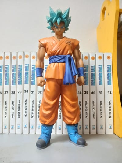 A full-body, slightly low-angle shot shows a Dragon Ball Z action figure with blue hair, orange gi, and blue belt standing on a white floor. The figure is positioned in front of a row of white manga volumes with "DRAGON BALL" and "AKIRA TORIYAMA" printed on them. A yellow, curved object is visible to the right of the figure. The background is a plain white wall. The image does not contain any information about the city of Monsummano Terme, Italy.