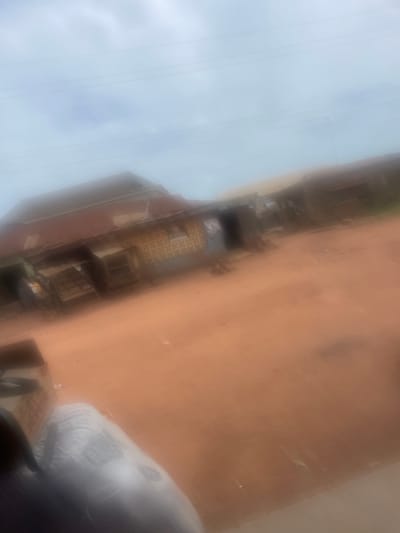 The image depicts a street scene in Ilero, Nigeria, characterized by a dusty, reddish-brown road. On the right side of the road, there are structures that appear to be small shops or homes with corrugated metal roofs. The buildings have patterned walls and some have open fronts or stalls. In the foreground, on the left, a large white sack or bag is visible, partially obscuring a cardboard box. The sky is overcast, suggesting cloudy weather. The overall impression is one of a typical day in a Nigerian town, with elements of commerce and everyday life.