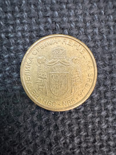 A close-up, eye-level shot captures a single, gold-toned coin resting on a textured, dark gray fabric surface. The fabric appears to be a woven material, possibly a rug or upholstery, with a coarse thread visible. The coin itself is round and features raised lettering and imagery.
The visible text on the coin reads "РЕПУБЛИКА СРБИЈА • NBS" which translates to "REPUBLIKA SRBIJA • NBS". "Republika Srbija" means "Republic of Serbia," and "NBS" likely refers to the National Bank of Serbia. The lettering is in Cyrillic script, with some Latin characters interspersed.
The coin's obverse displays a heraldic emblem, appearing to be a double-headed eagle with a crown, surrounded by intricate designs and what might be architectural elements. The overall condition of the coin suggests it has been handled, with some wear and possible minor discoloration, but the inscriptions and emblem remain legible.
The lighting in the image is somewhat diffused, highlighting the raised details of the coin and the texture of the fabric. There are no people or animals visible, and the focus is solely on the coin and its immediate surroundings. The image is static, depicting the coin at rest on the fabric. The location context provided is Mejaniga, Italy, but there are no specific visual cues within the image to confirm this location.