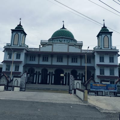 This full shot captures the exterior of a large mosque under an overcast sky. The mosque is a multi-story structure, predominantly white or light grey, with a distinctive green dome at its center, featuring a checkered pattern and topped with a crescent moon and star finial. Two tall, ornate minarets flank the main dome, each also crowned with a smaller green dome and a crescent moon and star. The building's facade is adorned with intricate geometric patterns and arched windows and entrances, reflecting Islamic architectural styles. Red-tiled overhangs provide contrast to the light-colored walls.
In the foreground, a paved road or courtyard leads up to the mosque, bordered by decorative white and brown fencing or gates. Utility wires stretch across the sky above the building. The lighting suggests it is daytime, but the cloudy conditions result in soft, diffused light.
On the right side, a blue banner is prominently displayed. The text on the banner reads: "DONASI WAKAF Untuk Pemasangan Keuremik Halaman Mesjid Sebelah Timur MASJID BESAR TAQWA LAMPOIH SAKA TAHUN 1447 HIJRIAH - 2026 MASEHI". It details donation goals, including "748 M X RP.230.000 - 748 M RP.172.040.000 -" and "KERAMIK YANG DIPASANG RP.100.000 KOTAK," along with contact phone numbers and bank details for "Bank Aceh." This indicates a fundraising campaign for the mosque, identified as "Masjid Besar Taqwa Lampoih Saka." The scene is located in Dua Paya, Indonesia.