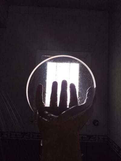 A hand, with fingers spread, is silhouetted against a bright, rectangular light source seen through a round, transparent object. The hand and the object appear to be in a dimly lit, tiled room, possibly a bathroom. There are faint streams of water or spray visible on the left side of the image, and the transparent object has small droplets or smudges on its surface. The background suggests a window or an opening with bars. The setting is likely a residential bathroom in New Bani Suef City, Egypt, given the context.