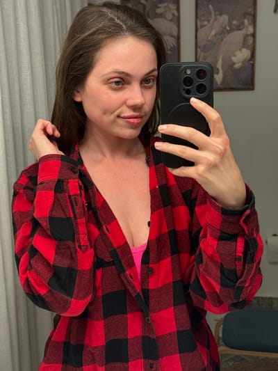 A woman is taking a selfie with a black smartphone. She is wearing a red and black flannel shirt, partially unbuttoned to reveal a pink top underneath. Her hair is brown, and she is looking directly at the camera with a slight smile. The background includes a wall with framed artwork and a blue chair, suggesting an indoor setting. This photo was not taken in Perugia, Italy.