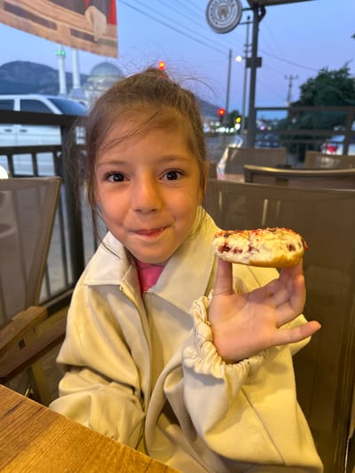 A young girl sits at a table, possibly outdoors, holding a sweet pastry with a red filling, perhaps in Köyceğiz, Turkey. The surrounding environment is somewhat blurred but suggests an urban area with streetlights and building lights visible in the background. She is wearing a beige jacket with gathered cuffs, and a pink garment underneath. The table has a wooden surface.