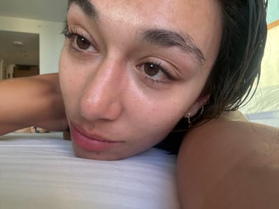 A close-up, slightly rotated view of a person's face, specifically the nose, eyes, and lips, is presented. The subject appears to be lying down, with a white textured surface visible above their mouth, possibly a pillow or bedding. The person has brown eyes, dark hair, and fair skin. A silver earring is visible on one ear. The background is blurry, but hints of a room can be seen, including part of a door and neutral-colored walls. It is not possible to determine the location as Honolulu based solely on the provided image.