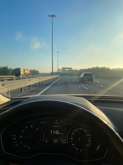 The image is taken from inside a car, looking out through the windshield at a highway. The car's dashboard is visible in the foreground, with a speedometer displaying a speed of 118 km/h. The road ahead is a multi-lane highway with a clear blue sky overhead, and some wispy clouds. There are tall streetlights along the highway. In the distance, a truck is visible on the left lane, and a dark SUV is in the right lane. The sun is low in the sky, casting a warm glow and creating lens flares. The scene is set on a highway in the vicinity of Zanevka, Russia, as indicated by the on-screen text. No people are visible in the image.