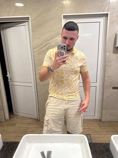 A young man with a short haircut and a beard takes a selfie in a restroom. He is wearing a yellow polo shirt with a white floral pattern and beige cargo shorts. He is holding a smartphone in his right hand. The restroom has beige tiled walls and wooden floor tiles. There are two white doors visible in the background. In the foreground, a white sink is visible. The image does not provide any information that allows for a location in the city of Gelemenovo, Bulgaria.