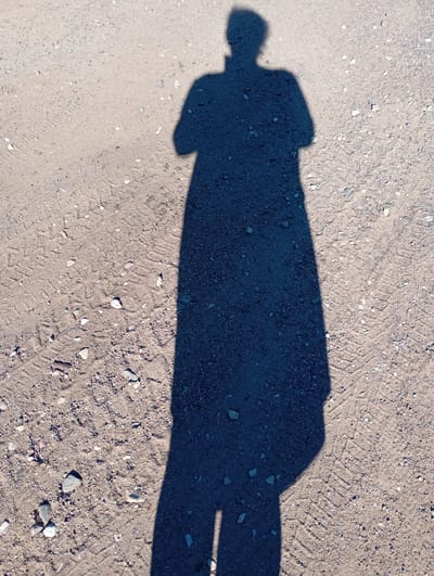 The image displays the shadow of a person standing on a dirt road. The shadow is long, suggesting it was taken during the early morning or late afternoon, when the sun is low in the sky. The ground is composed of sand and small pebbles, with tire tracks visible, indicating it's a road or path used by vehicles. The overall scene is simple and devoid of other subjects or objects. The lighting and the absence of any other cues prevent the determination of specific weather conditions or emotions. There is no visible text in the image. The location context provided is Ретюнь, Russia, which helps to understand the general geographic area, but offers no specific visual information within the image itself.