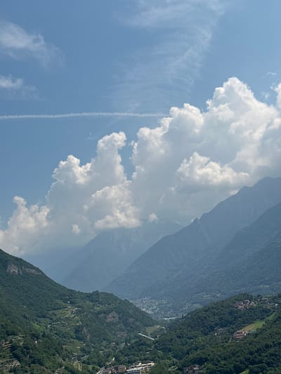 This media file depicts a scenic view of a mountainous landscape under a partly cloudy sky. The scene is dominated by lush green mountainsides dotted with buildings, suggesting a populated area within a natural setting. The slopes are steep and covered in dense vegetation, with some visible terraces or cultivated areas on the mountainside. A clear sky with wispy clouds and a prominent, large cumulus cloud formation is visible on the left side of the frame, contrasting with the darker, hazy mountain ranges in the distance. A contrail from an aircraft streaks across the sky, indicating possible air traffic.

The time of day appears to be daytime, likely late morning or early afternoon, given the brightness of the scene and the appearance of the clouds. The weather seems fair, with a mix of sun and clouds. The location cues, such as the architecture of the buildings visible on the mountainside and the mountainous terrain, are consistent with a location like Malegno, Italy. No people or specific objects are clearly discernible in the foreground or mid-ground, making the focus primarily on the grand natural scenery and the human settlements integrated within it. There is no visible text in the image. The overall impression is one of natural beauty combined with human habitation in a mountainous region.