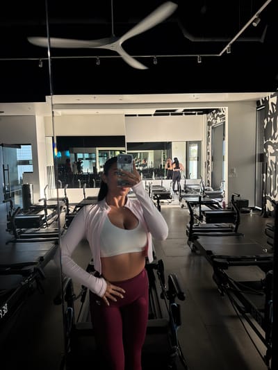 A woman in a white sports bra and leggings poses in front of a mirror at a gym in Long Beach, California. The gym has rowing machines and is decorated with black and white patterns on the ceiling. Other people are working out in the background. The woman is holding a phone and taking a selfie.