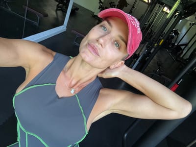 An outdoor, selfie-style, medium shot shows a woman posing in a gym. The woman, who appears to be in her late 30s or early 40s, has light skin, freckles, and green eyes. She is wearing a pink baseball cap and a dark gray workout tank top with neon green stitching. Her hair is light brown, and she is wearing a silver necklace with a heart-shaped pendant. She is leaning her head on her right arm, with her left arm bent upwards and resting near her head. The background shows gym equipment, including weight machines and benches, and a mirror reflecting a portion of the gym. The overall setting suggests a private or semi-private gym space. The location is identified as Ivanovići, Montenegro.