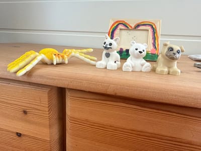 A yellow and orange plush spider toy lies on its side on a wooden surface. To the right of the spider, there are three plastic figurines: a white cat, a white bear, and a tan pug. Behind the figurines, a wooden picture frame with a colorful rainbow drawing inside is leaning against a white wall. The wooden surface appears to be the top of a dresser or desk, with visible wood grain and knots. The scene is indoors, likely in a domestic setting. There are no people visible in the image.
