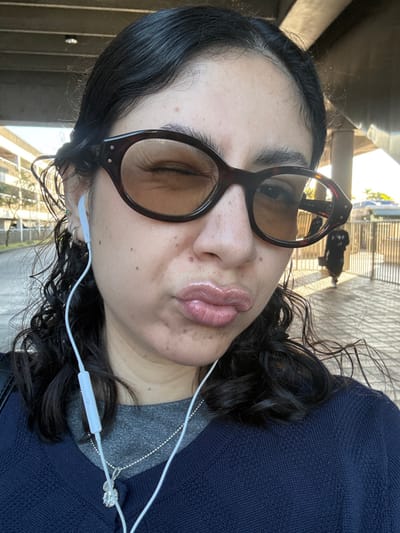 A front-facing, waist-up image captures an adult female with dark, wavy hair, illuminated by natural light in an outdoor urban setting. She wears tortoise-shell oval sunglasses with light amber lenses; her right eye is visibly closed in a wink, creating wrinkles at the outer corner. Her lips are pursed, appearing glossy with a light pink or nude product. White wired earphones are in both ears, with an inline control visible on the wire resting on her chest. Her right ear features multiple piercings, including a stud with a green stone and several silver rings. She is dressed in a dark blue v-neck sweater over a grey shirt. A silver ball-chain necklace with a circular, clear faceted pendant hangs around her neck. In the background, a large, grey concrete elevated structure dominates the upper frame. To the left, a multi-story building is partially visible. To the right, a blurry, dark silhouette of another individual walks away behind a vertical-barred metal fence. The environment is consistent with urban infrastructure, such as elevated transit lines or roadways, within the city of Hialeah, Florida, United States of America.