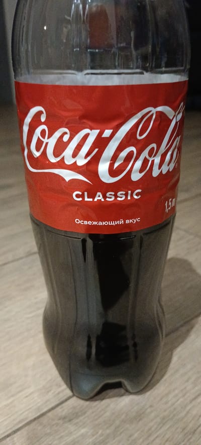 A clear plastic bottle of Coca-Cola Classic, 1.5 liters, is standing upright on a wooden floor. The bottle is partially full of dark liquid. The label is red with white text, featuring the Coca-Cola logo, the word "CLASSIC," and the phrase "Освежающий вкус" (Refreshing taste) in Russian. To the right of the "CLASSIC" text, the volume "1,5 л" is visible. The background is dark and out of focus. The image is taken from a slightly low angle, focusing on the bottle. The location in the city of Падлабенскі сельскі Савет, Belarus, is not depicted or inferable from the image itself.